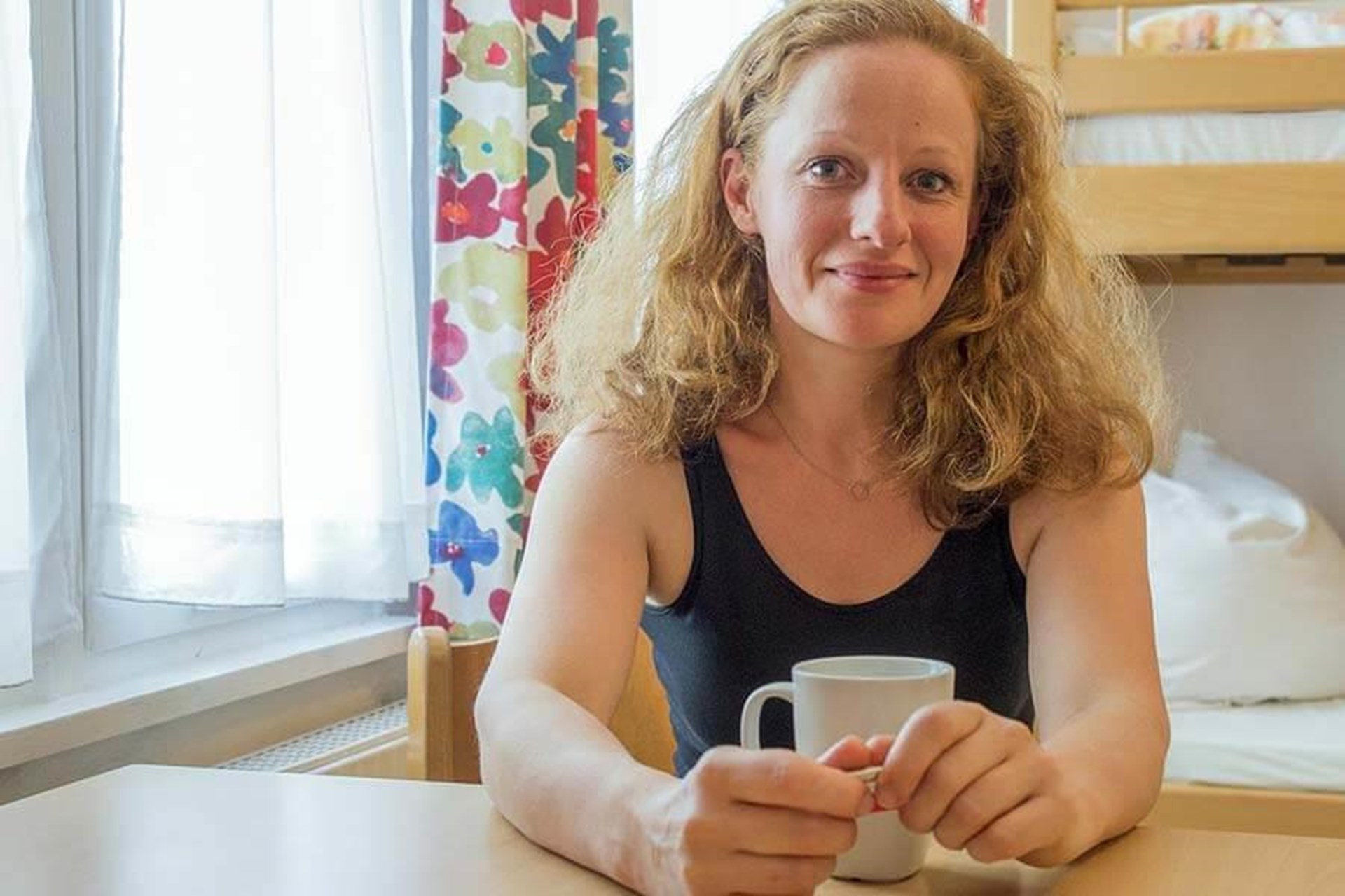 Frau mit blondem, lockigem Haar sitzt an einem Tisch und hält eine weiße Tasse, im Hintergrund sind bunte Vorhänge und ein Fenster zu sehen.