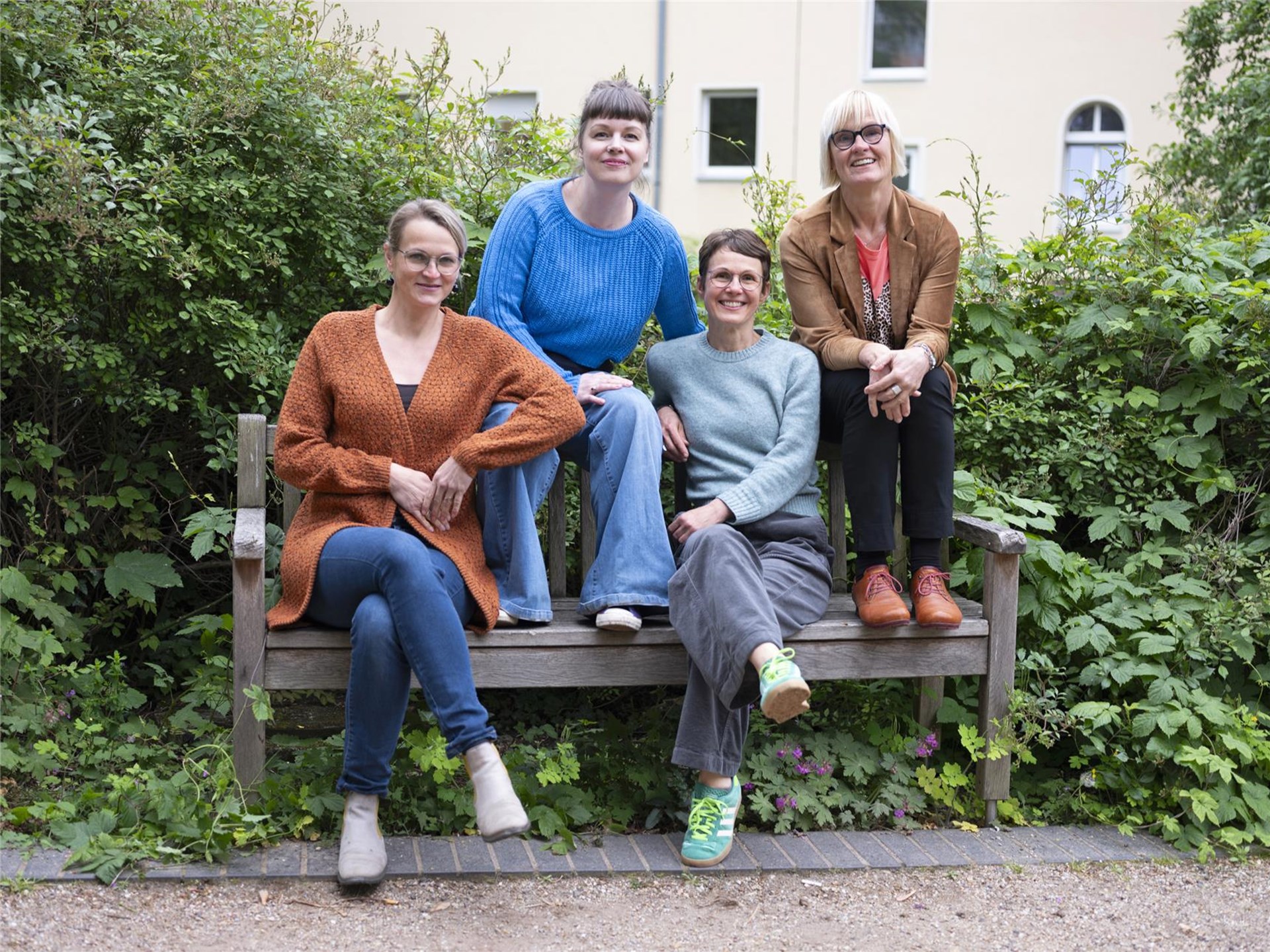 Gruppe von vier Frauen sitzen auf einer Bank und schauen freundlich in die Kamera. Sie sitzen vor einem grünen Gebüsch.