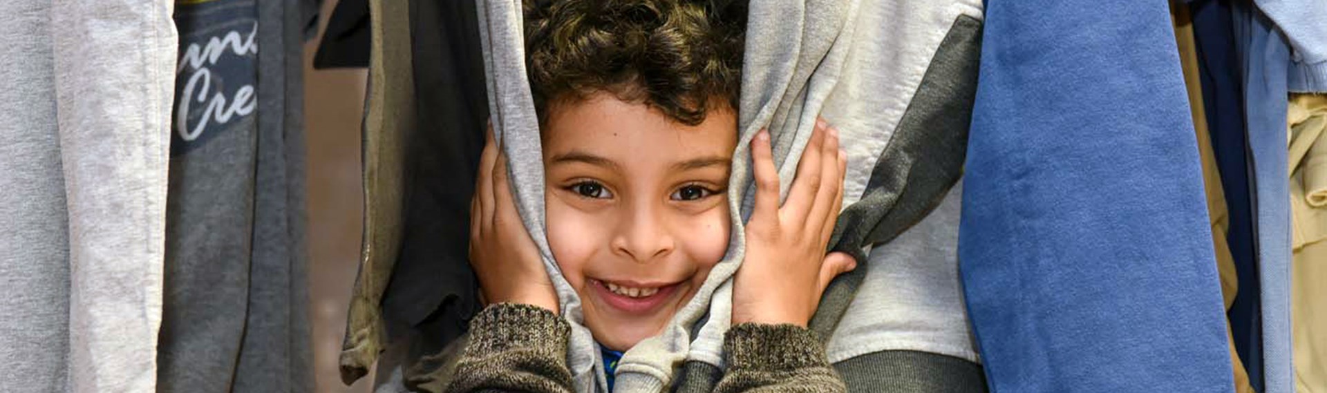 Portrait eines kleinen Jungen, der sein Gesicht zwischen den Klamotten eines Kleiderständers hervorstreckt und lacht