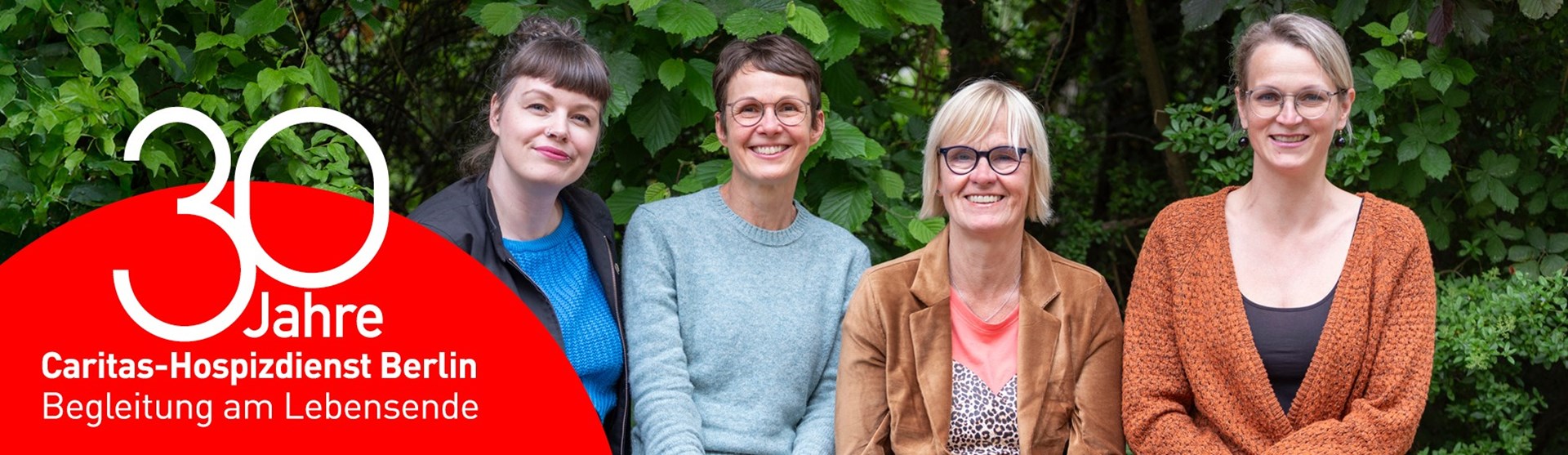 Vier Frauen sitzen lächelnd vor grünem Laub. Links im Bild steht in rotem Kreis: „30 Jahre Caritas-Hospizdienst Berlin – Begleitung am Lebensende“.